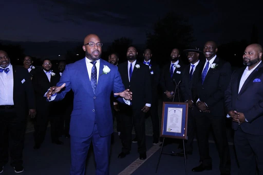 Tau Zeta Sigma brothers in suits gathered outdoors at night beside the chapter charter